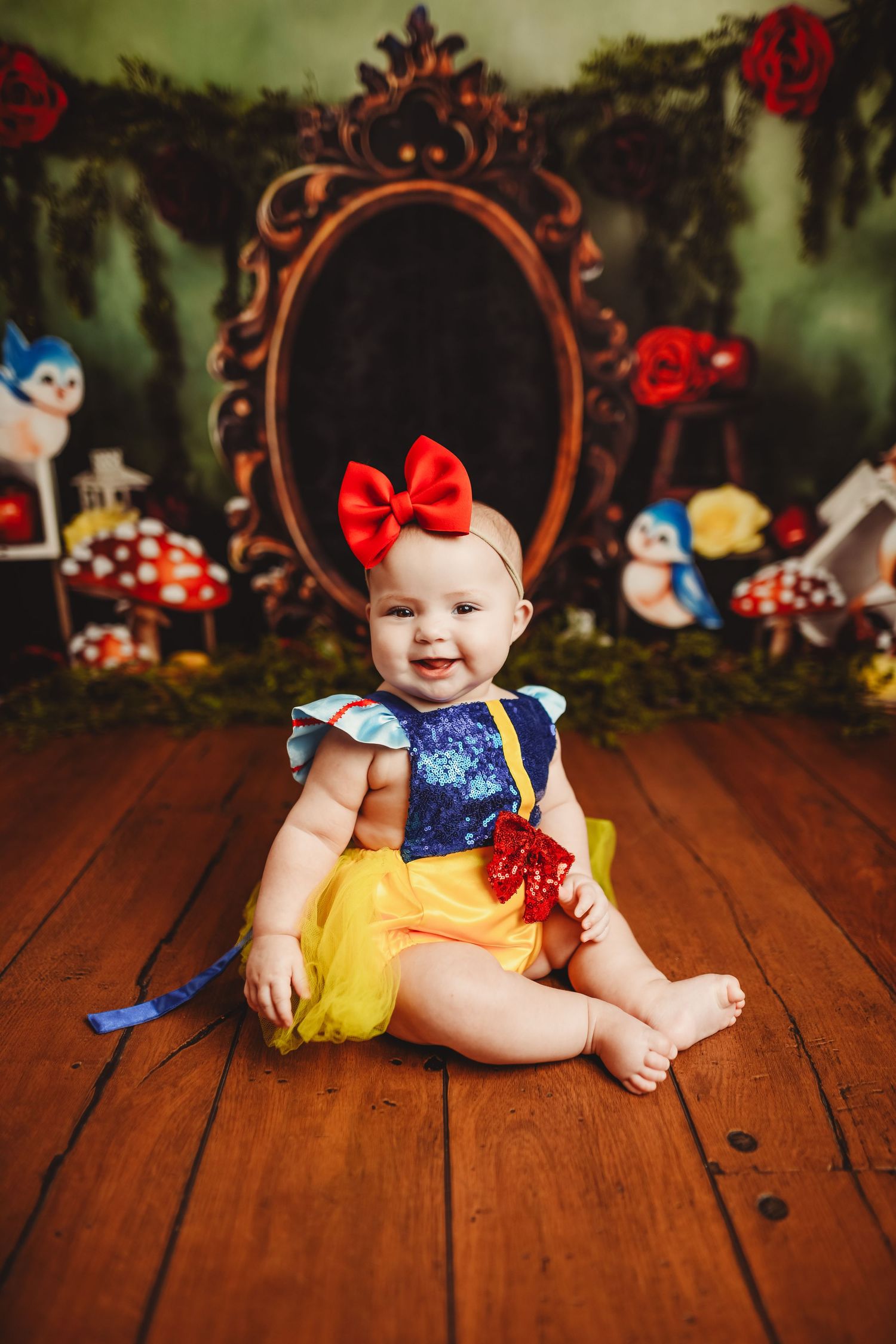 Baby in Snow White inspired costume on wooden floor with enchanted forest backdrop.
