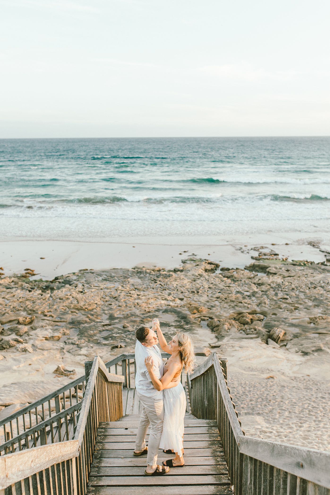 A couple shares a romantic moment on wooden stairs overlooking a coastal beach.