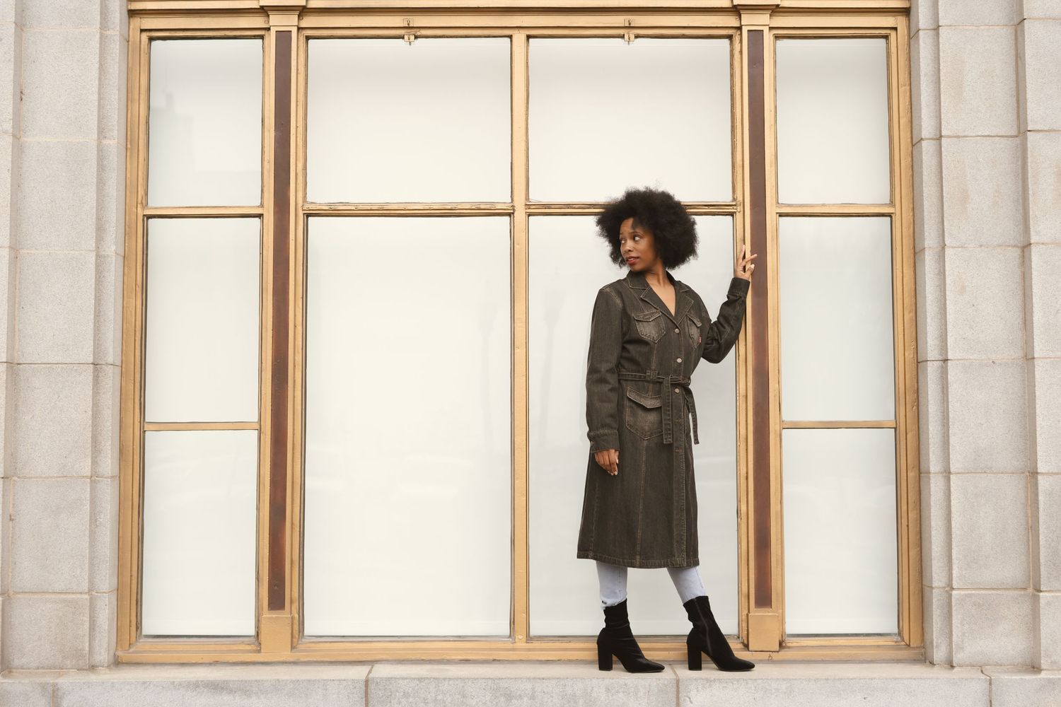 a woman in a trench coat standing on a large outdoor concrete window frame and looking behind her