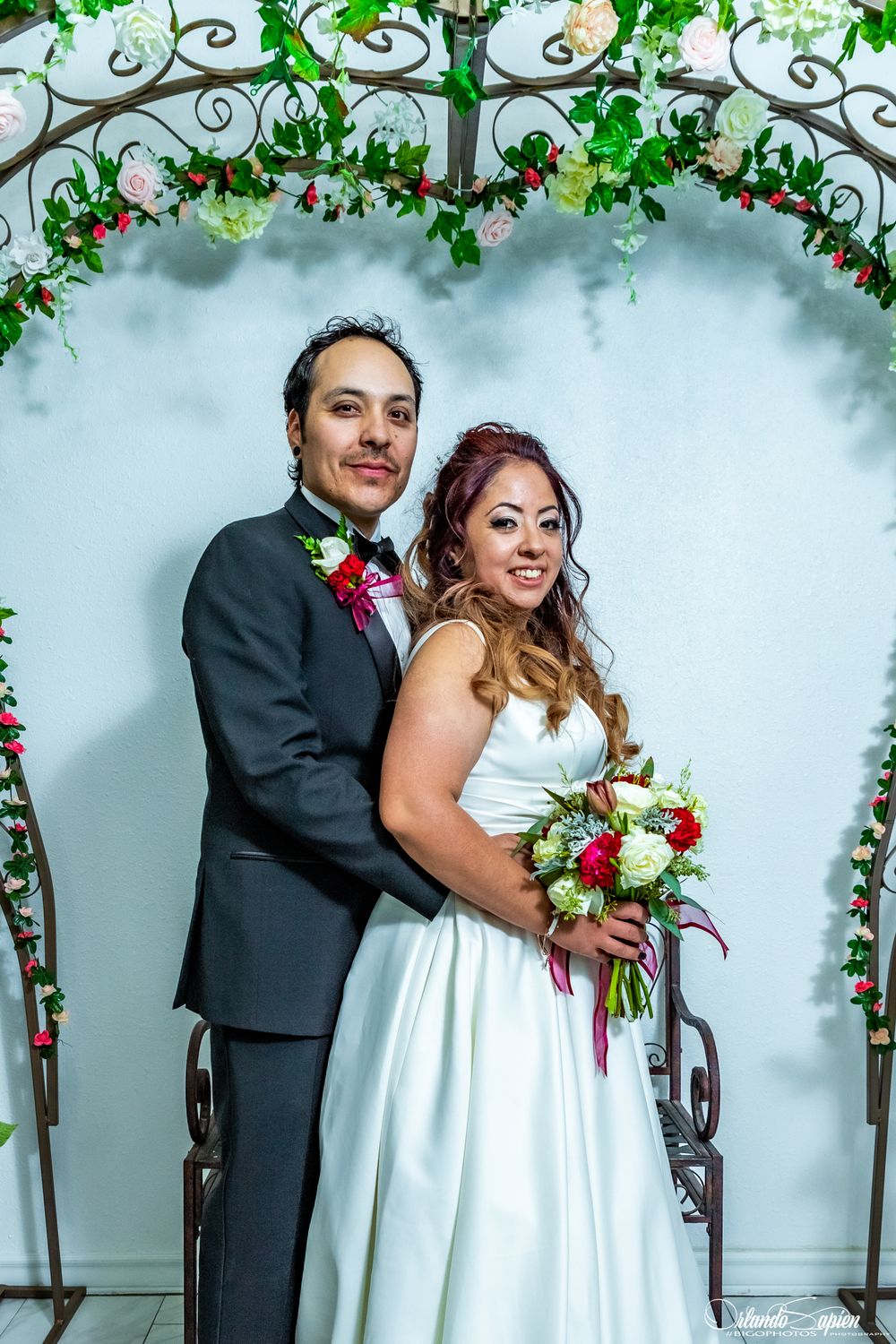 Wedding couple poses under floral arch with decorative vines and flowers.