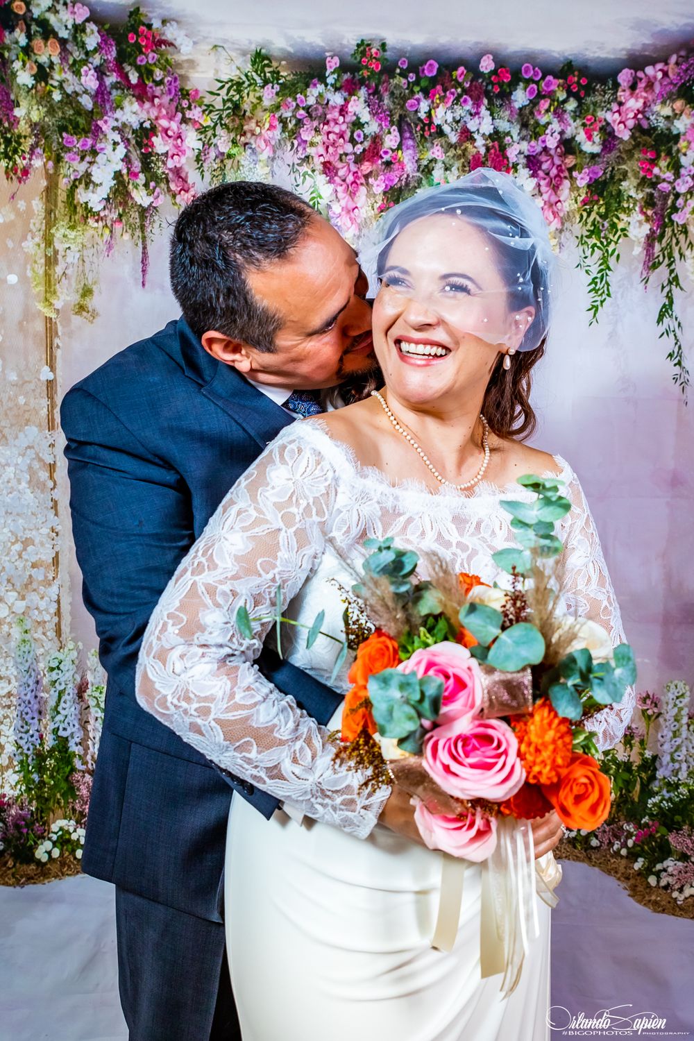Bride and groom share intimate moment under colorful floral canopy.
