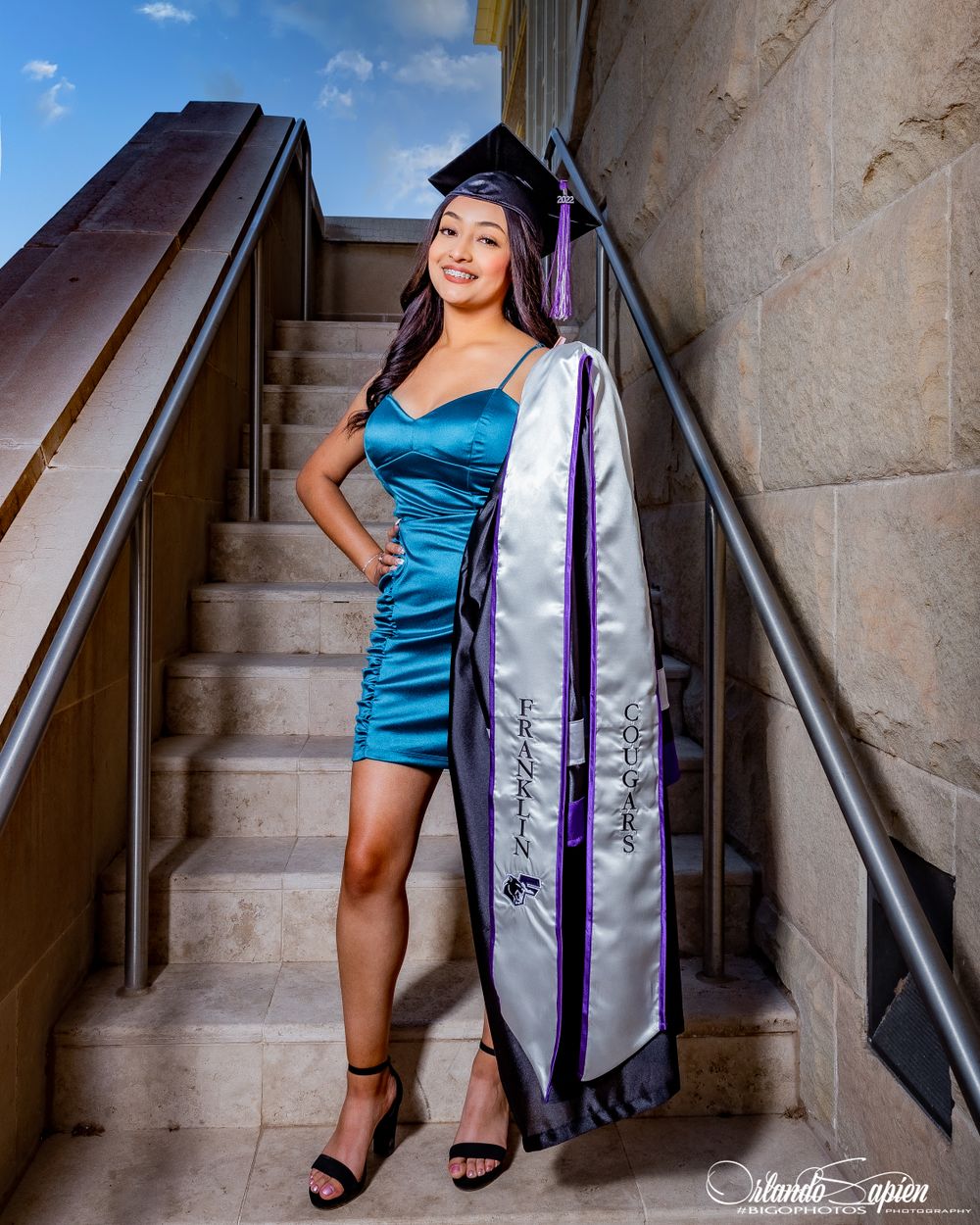 Graduate in teal dress and purple stole poses on concrete stairs for graduation photos.