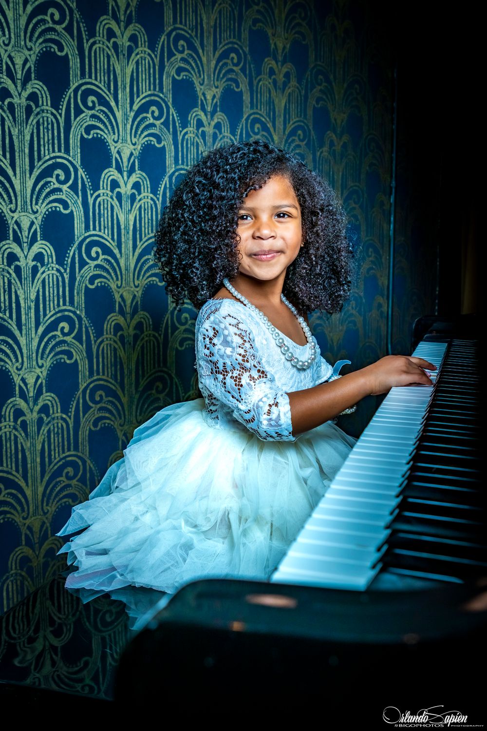 Young pianist in blue tulle dress performs at elegant piano against art deco wallpaper.