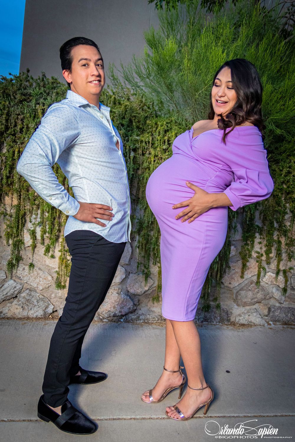 Maternity photo of couple in casual wear posing playfully against garden wall.