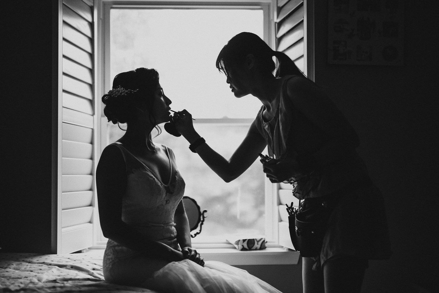 A silhouetted figure applies makeup to a seated person by a window in black and white.