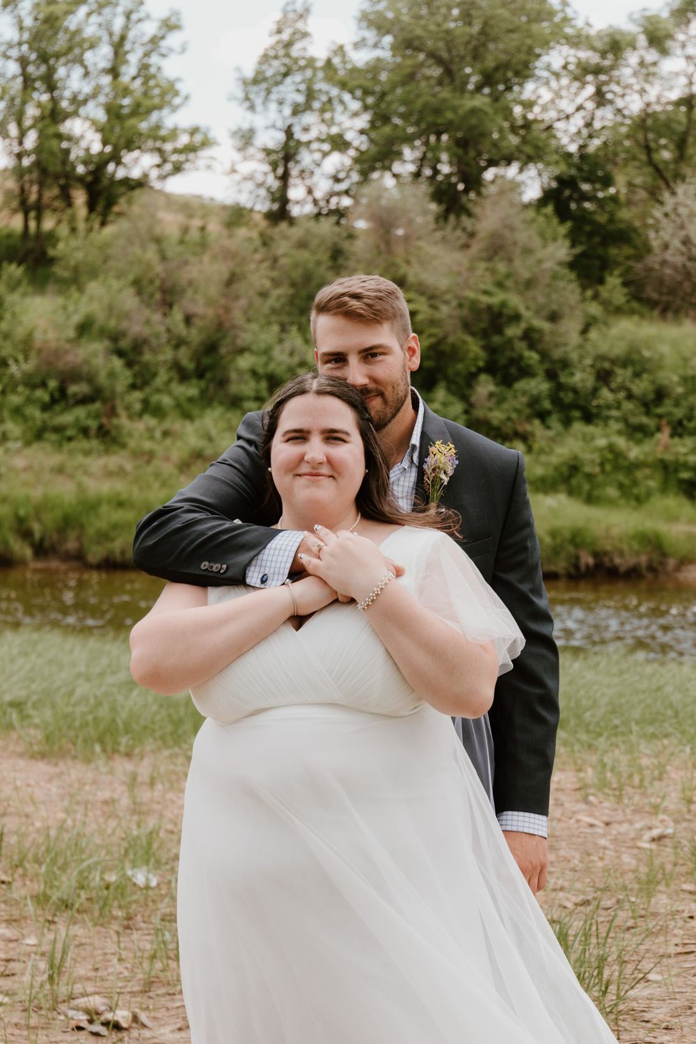 Newlyweds share a tender moment by a river with lush greenery in background.