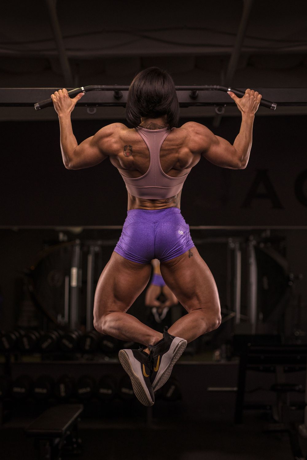 Muscular back view of athlete performing pull-up in purple shorts at gym.