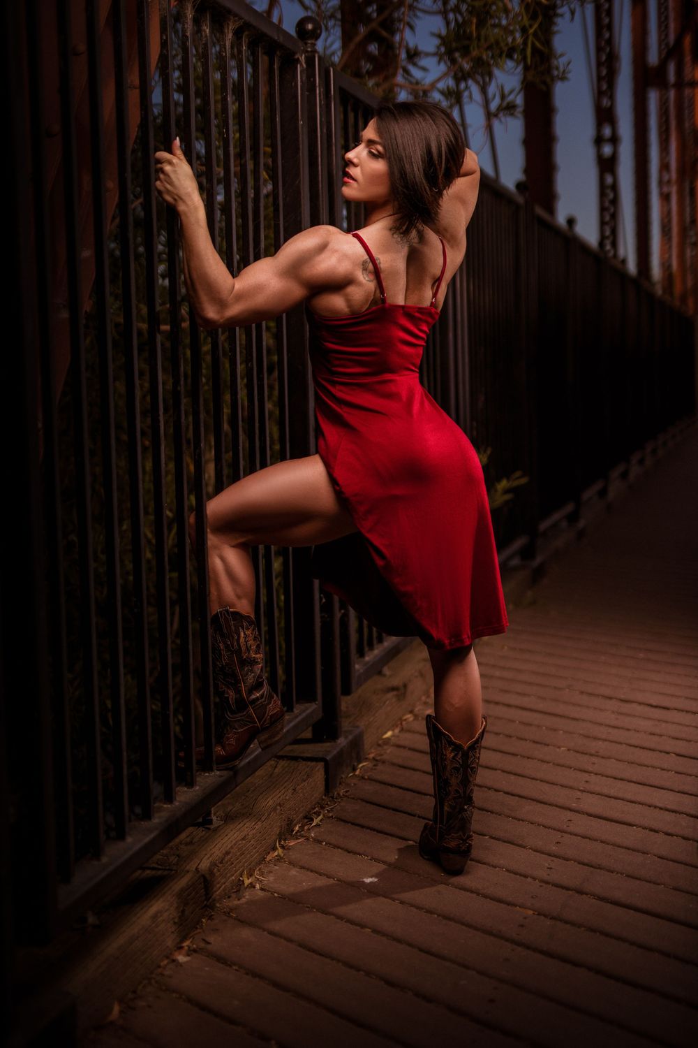 Athletic figure in red dress shows strength while posing on railing at dusk.