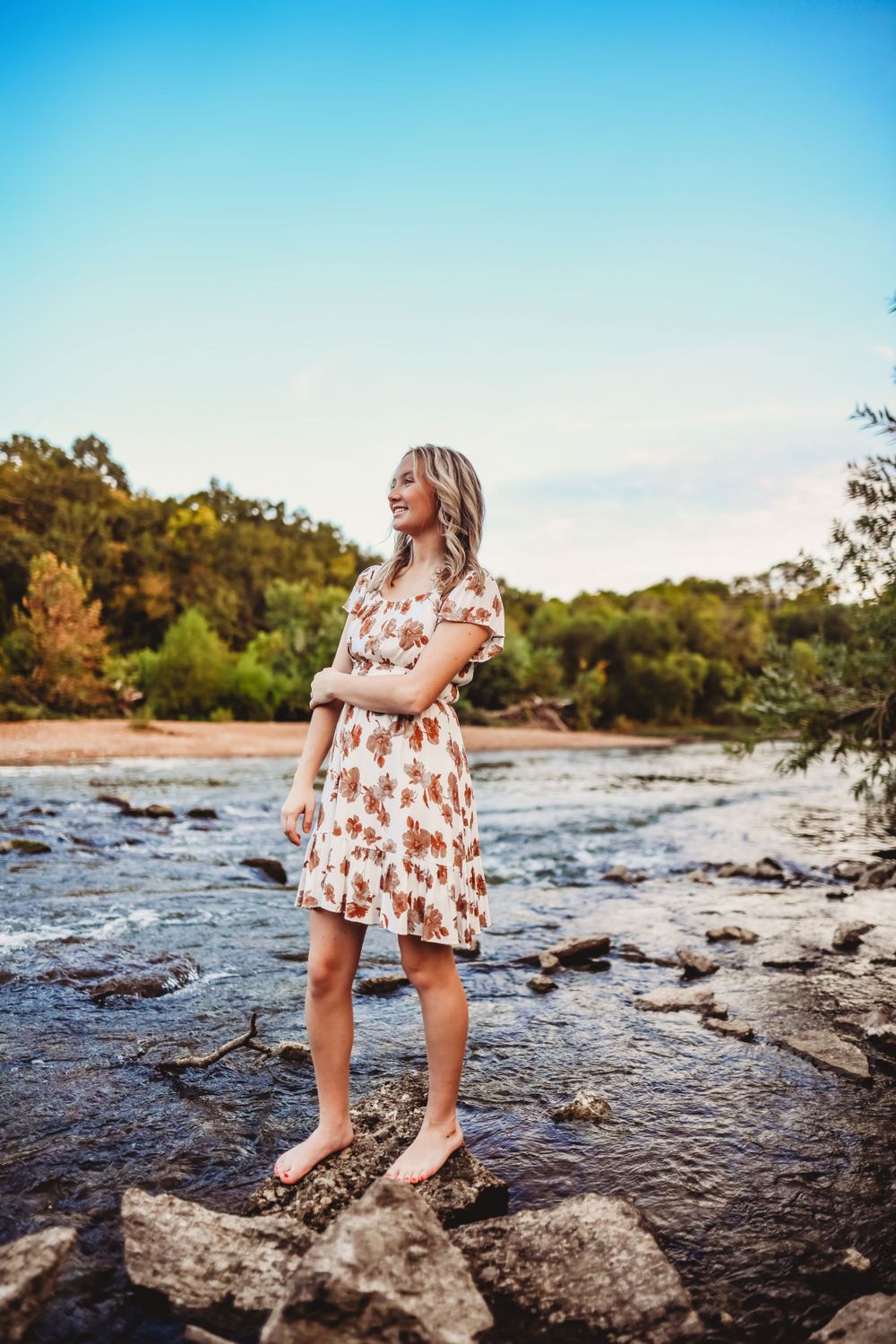 White dress with brown pattern stands on rocks by flowing river under blue sky.