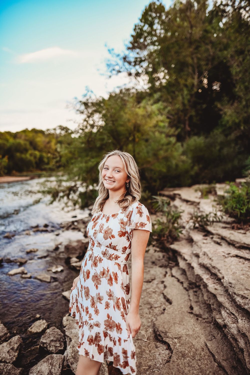 Floral dress stands on rocky riverbank against blue sky and tree backdrop.
