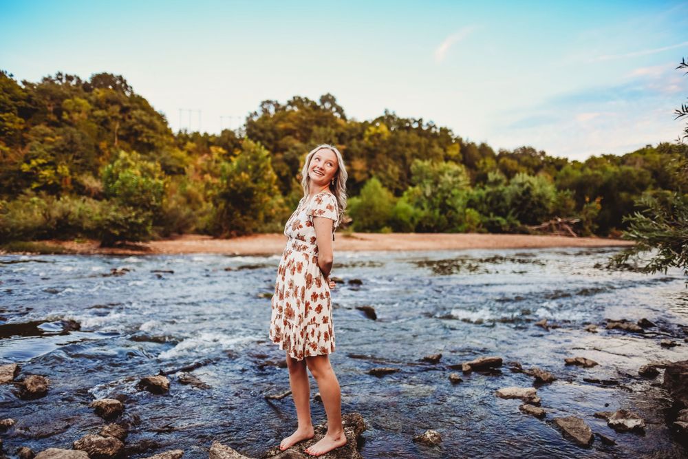 Floral dress poses gracefully on rocky riverbed with water flowing behind.