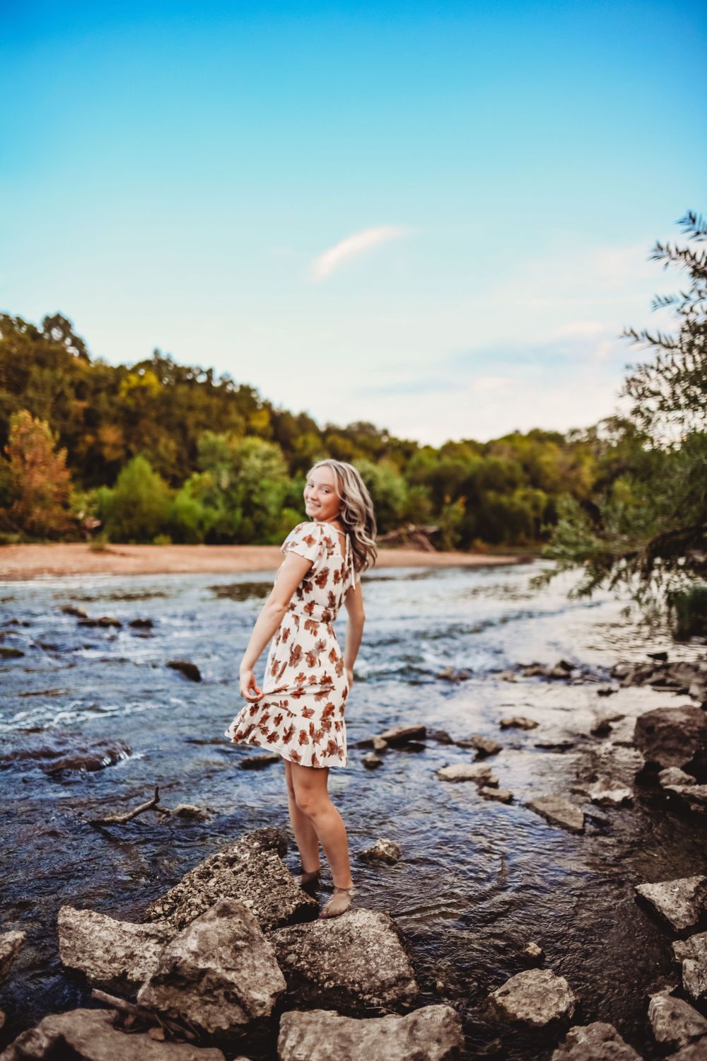 Floral dress balances on river rocks with flowing water and autumn trees in background.