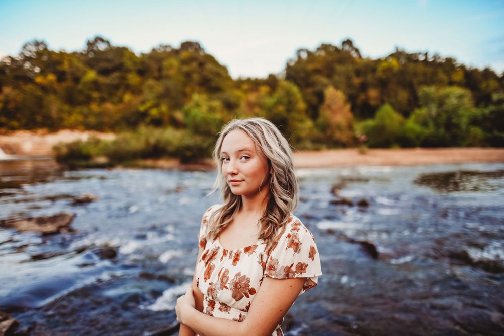 Floral dress stands against backdrop of rushing river and autumn foliage.