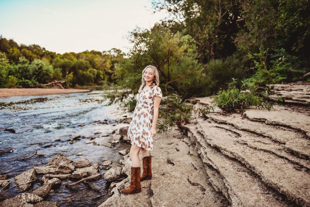 Brown boots stand on rocky riverbank in white floral dress during sunset.