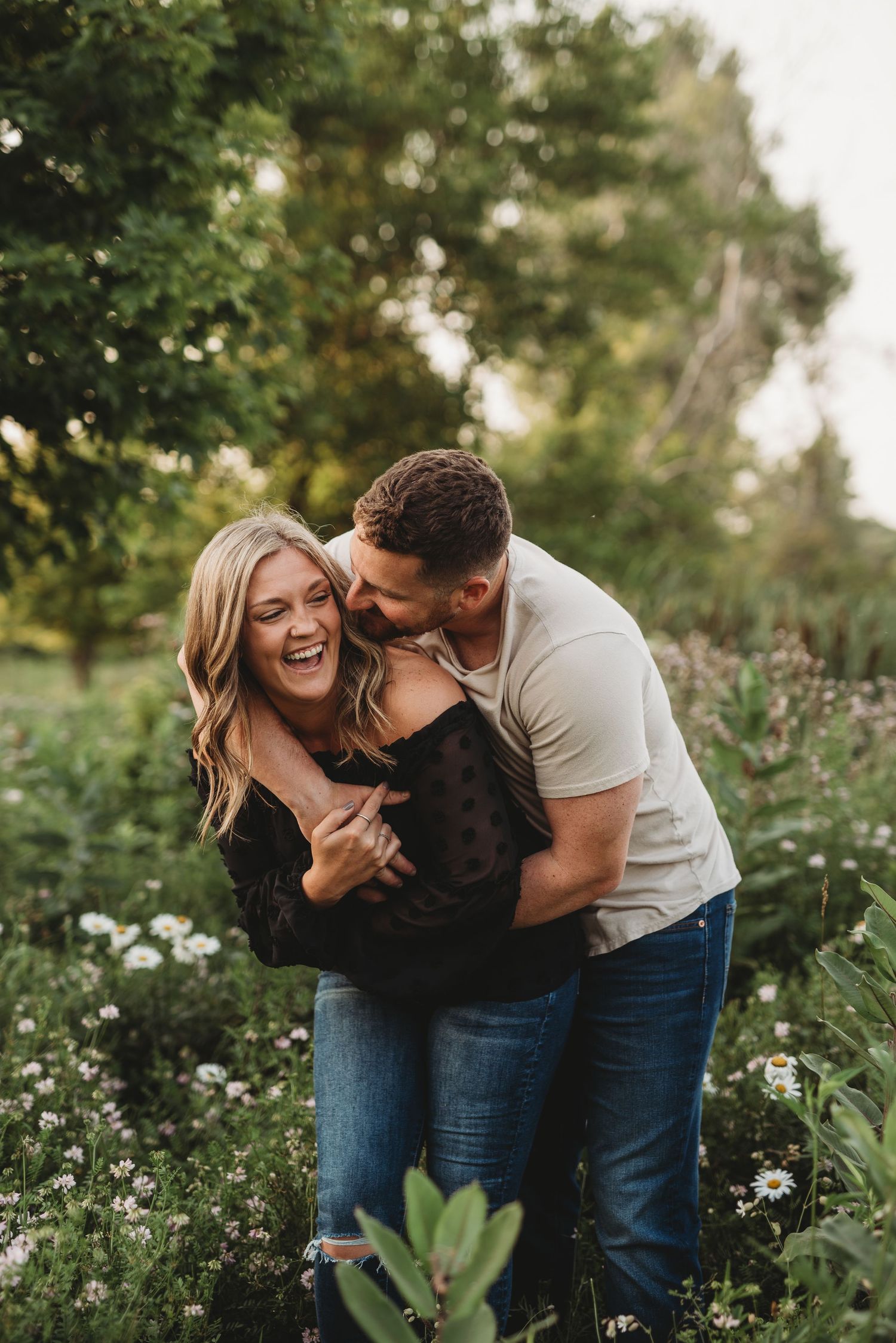 A couple shares a joyful embrace among wildflowers and greenery in a natural outdoor setting.