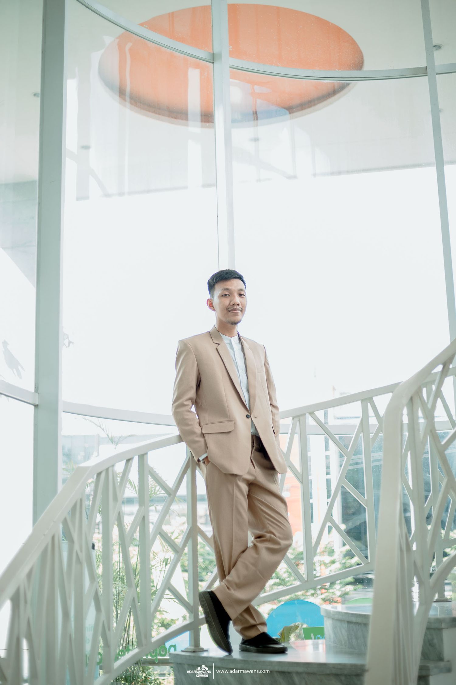 Groom in beige suit poses confidently on modern spiral staircase with orange ceiling detail.
