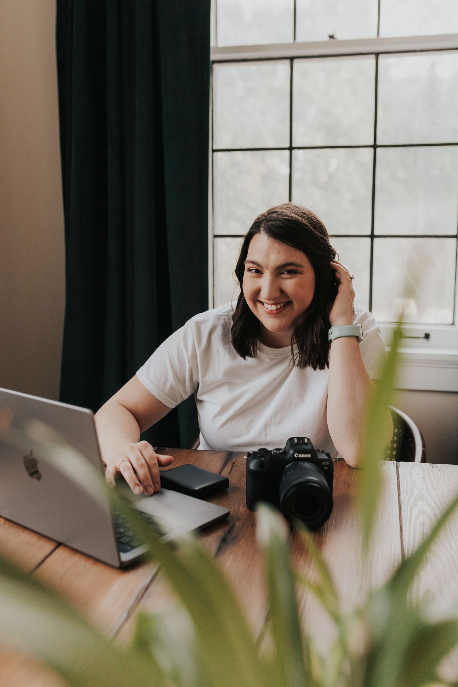 Content creator working at desk with laptop and camera near window with plants.