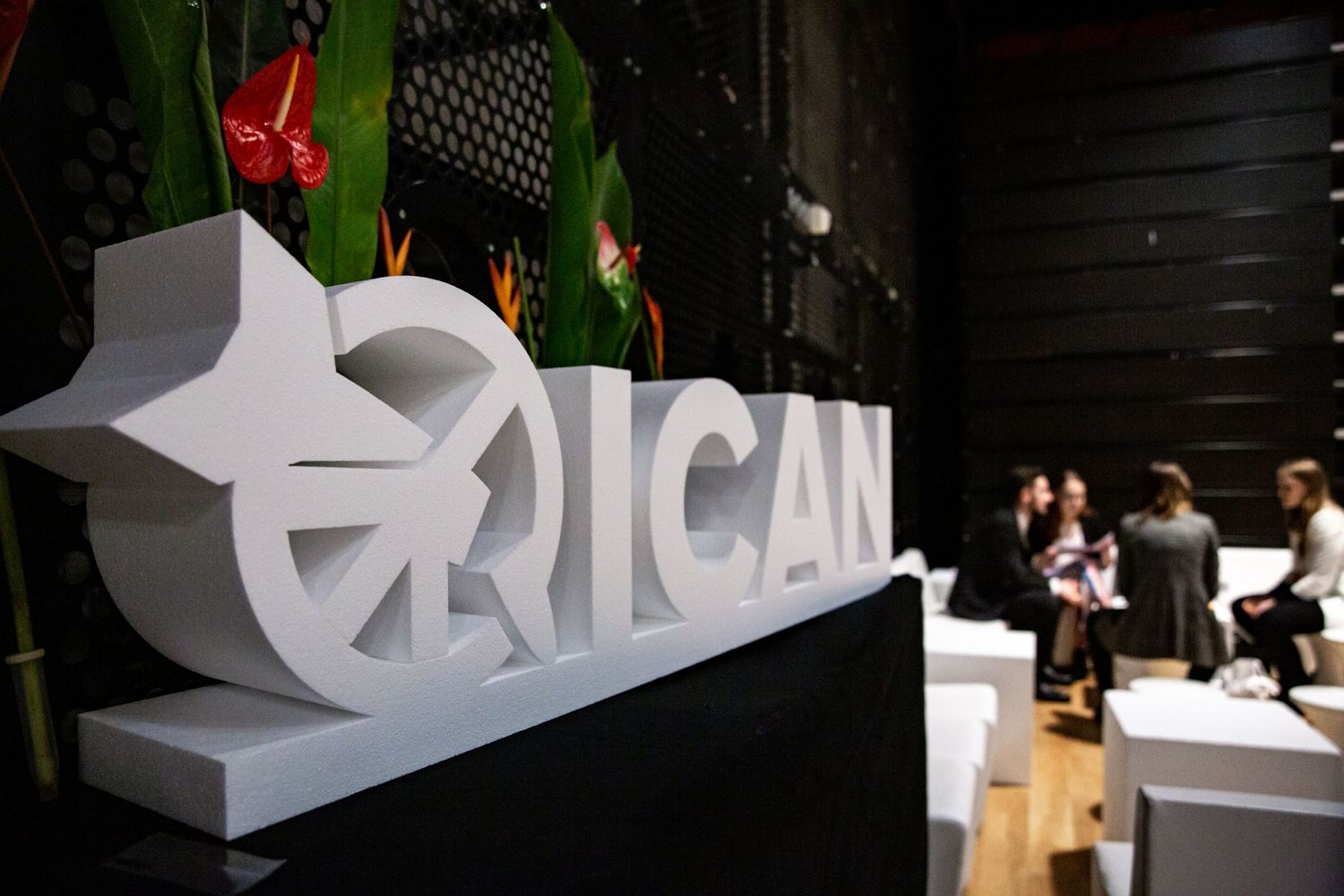 Large white ICAN sign displayed at an event venue with people gathering in the background.