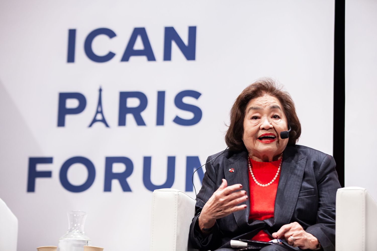 Speaker at ICAN Paris Forum event against branded backdrop with organization logo.
