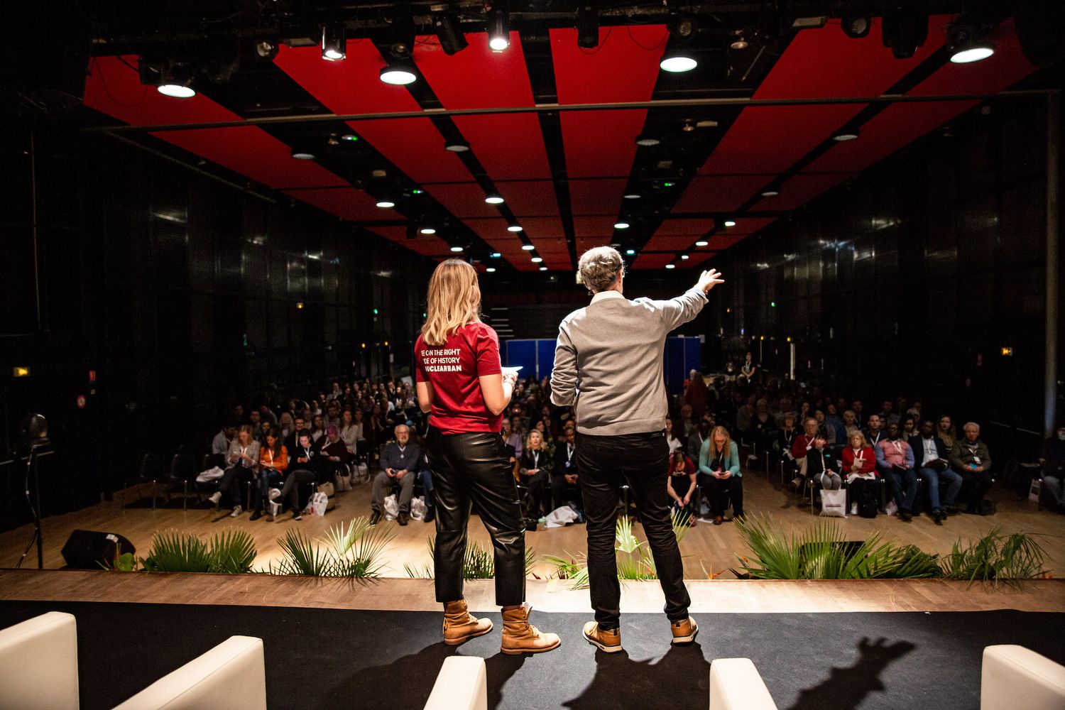 Two presenters speaking on stage to audience at conference with red ceiling lights.