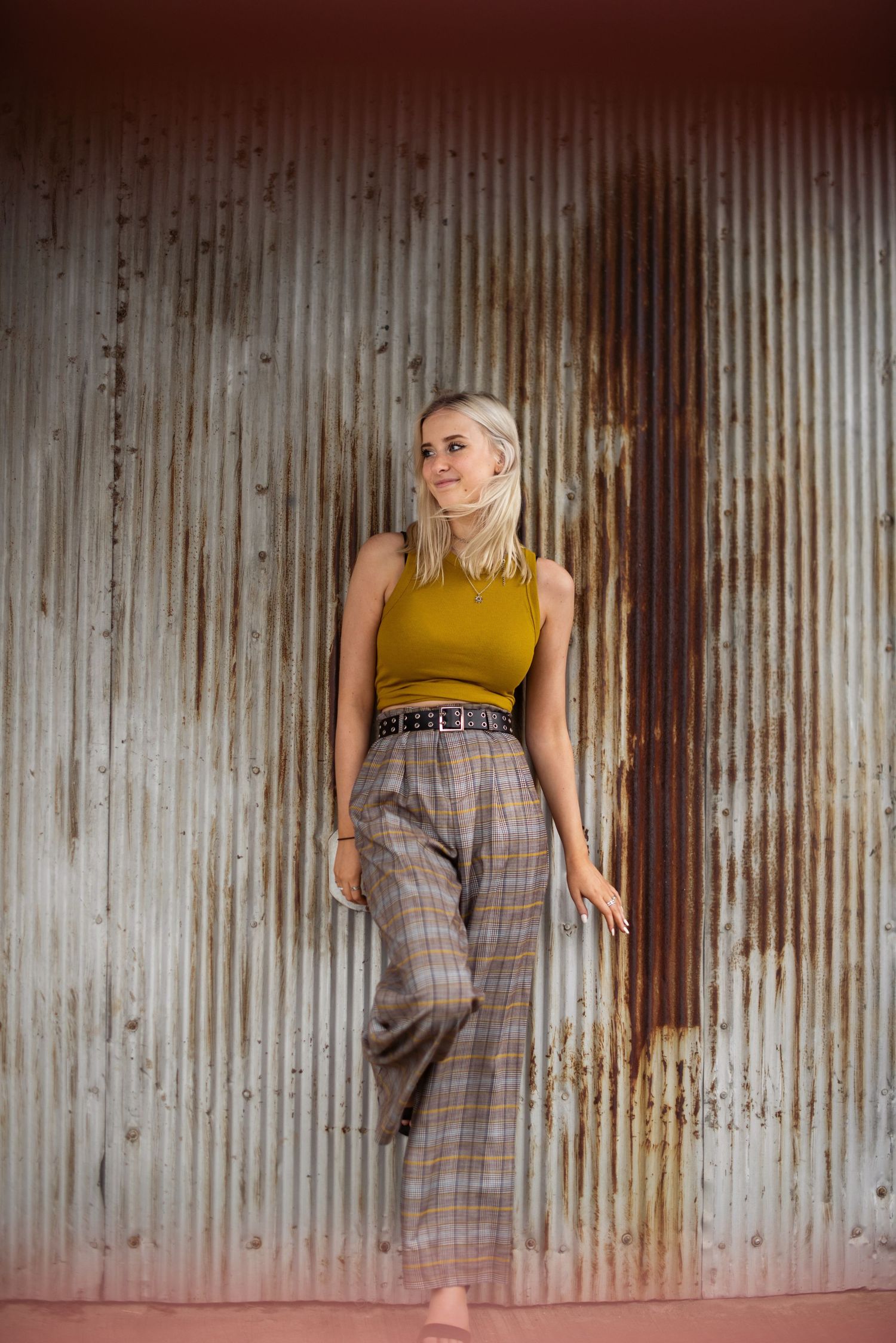 Fashion portrait showing person in yellow top and plaid pants against corrugated metal wall.