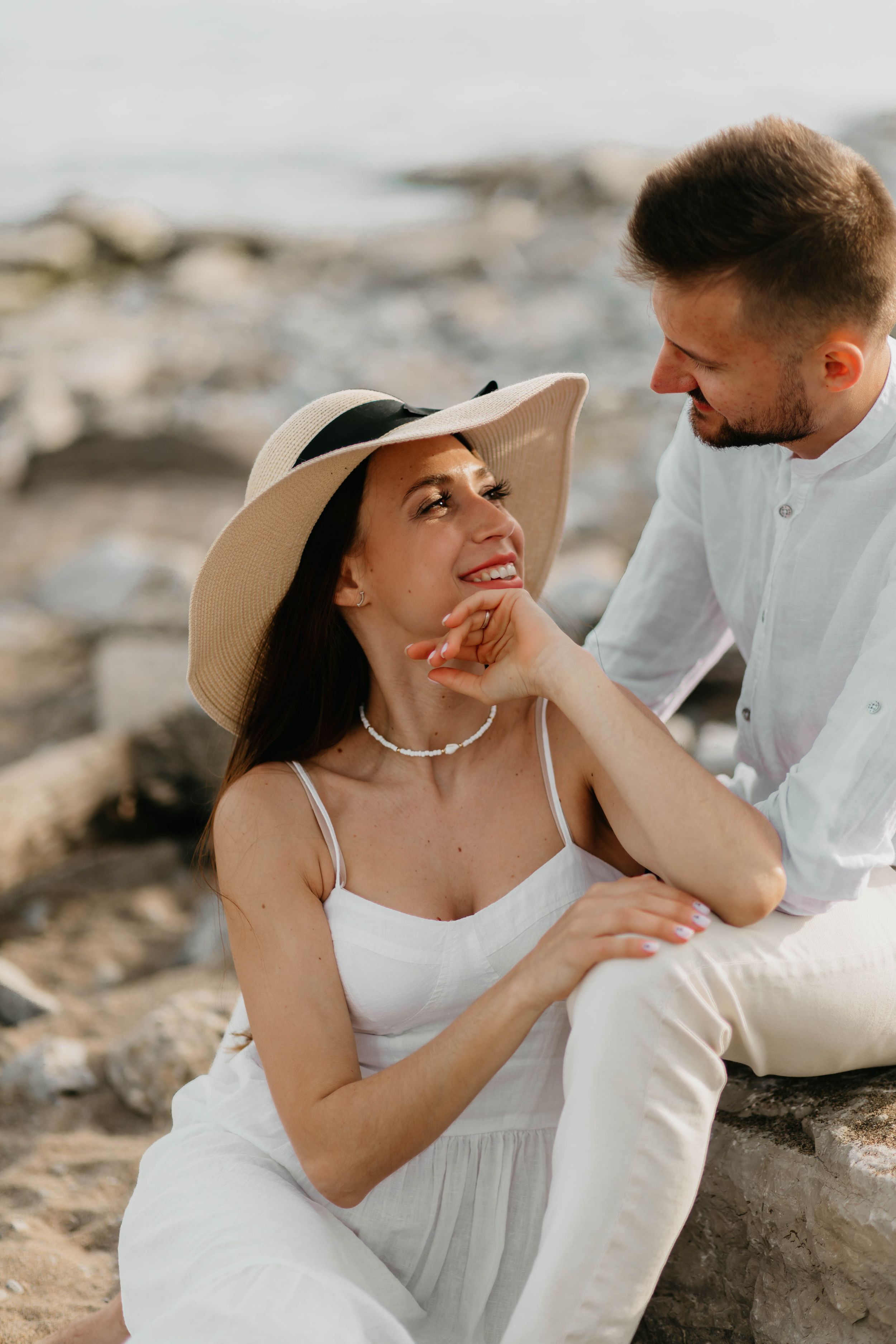 Couple in matching white outfits share tender moment on rocky coastline.
