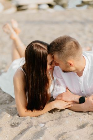 Romantic couple lying on sandy beach sharing an intimate moment in white outfits.