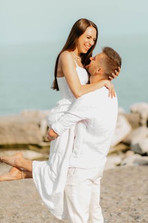 Joyful couple in white embracing on rocky beach during sunset photoshoot.