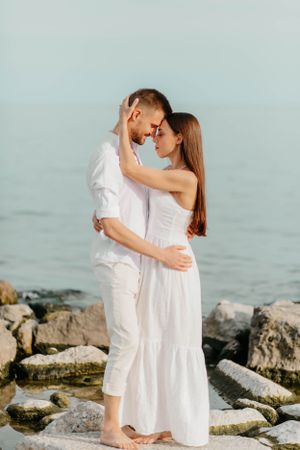 Couple in all white embracing on rocky shoreline against misty ocean backdrop.