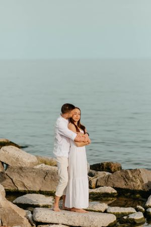 Couple standing on coastal rocks sharing embrace against serene ocean view.