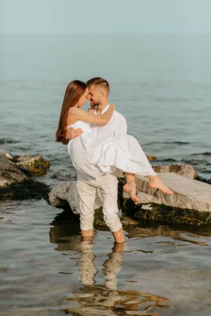 A couple in white embracing while standing in shallow beach water near rocks.