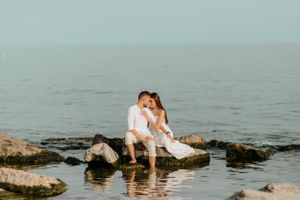Couple in white outfits sitting together on rocks by the water's edge.
