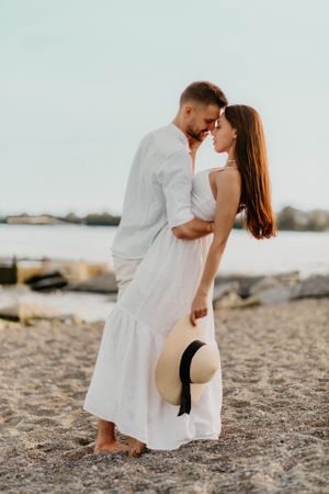 Couple in white outfits embracing on beach, woman holding straw hat.