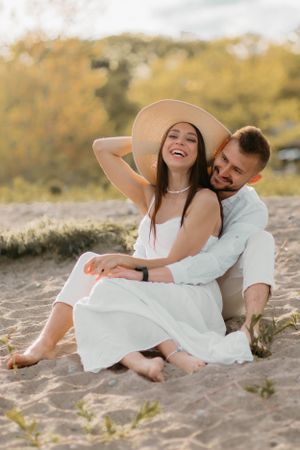 Happy couple in white relaxing on sandy beach, woman wearing straw hat.
