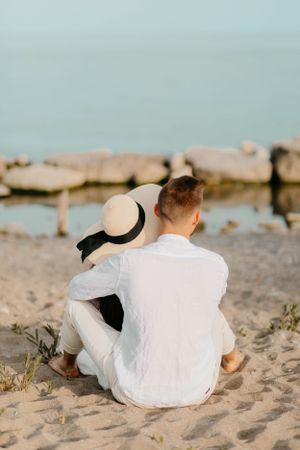Person in white shirt sitting on beach with straw hat watching the ocean.