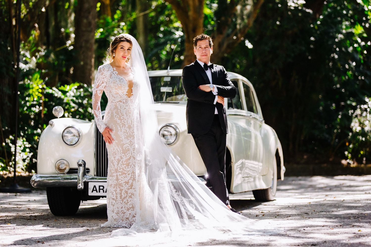A bride and groom walk along a lush tree-lined pathway in sunlight.