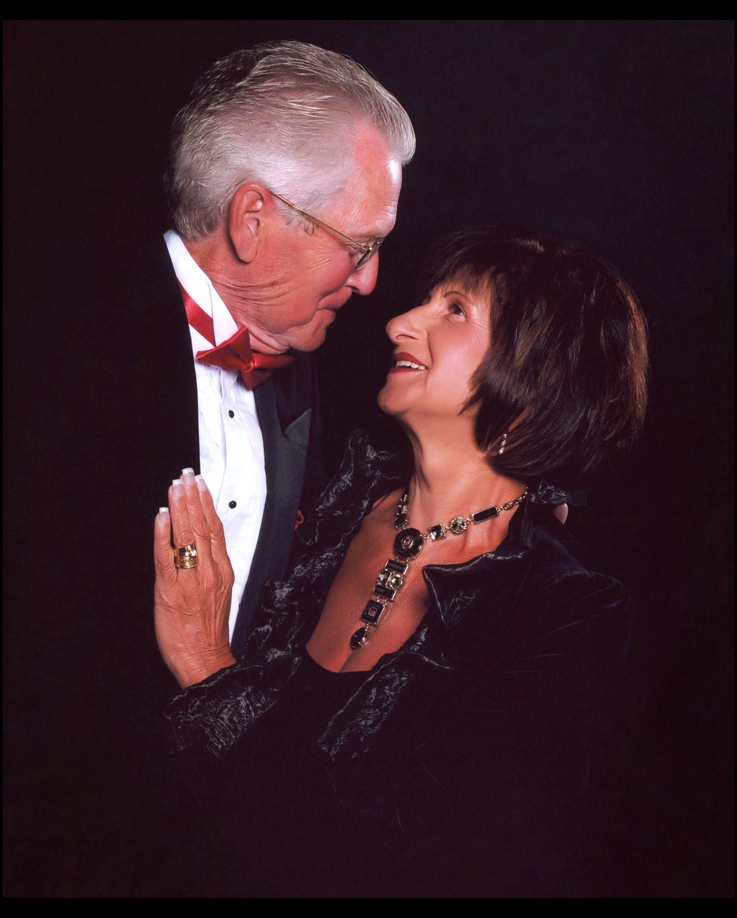 Formal couple embracing and smiling in evening wear against dark background.