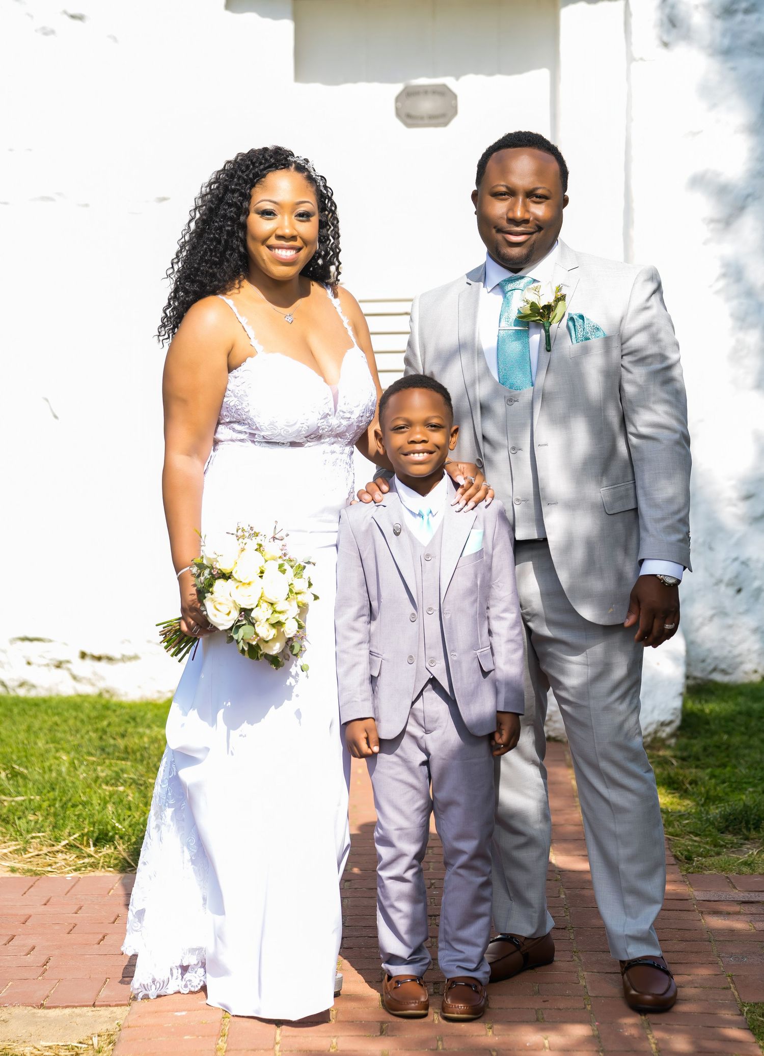 A wedding party poses together in formal attire outside a white building.