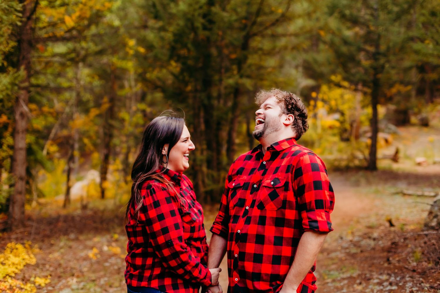 Couple wearing matching red plaid shirts laughing together in a fall forest setting.