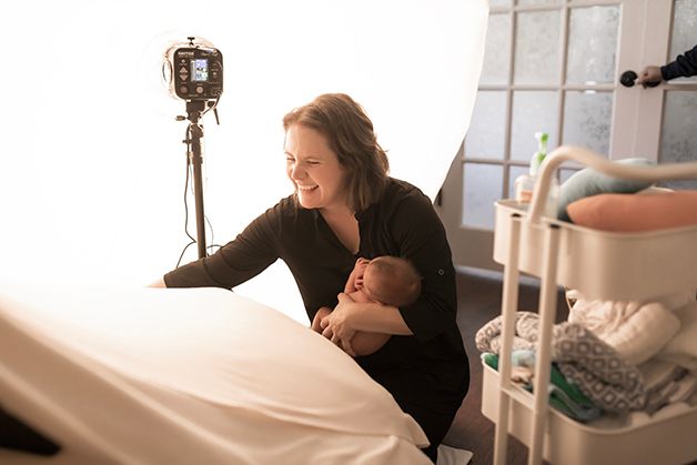Laura sitting and holding newborn baby laughing while securing blankets for a pose shot.