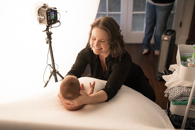 Laura, owner of Hope Believe Photography, smiling holding a newborn baby posing him on beanbag.