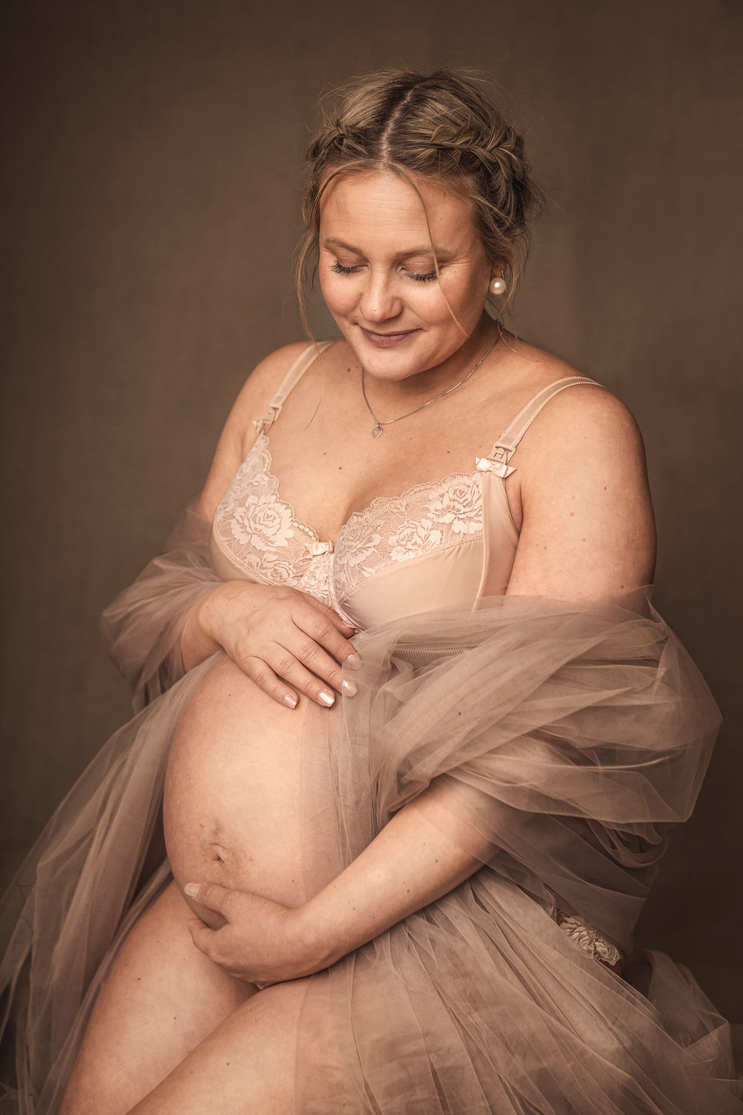 Gravid kvinne på mørk beige bakgrunn i undertøy med tyll rundt seg som holder på magen og ser ned på den og smiler