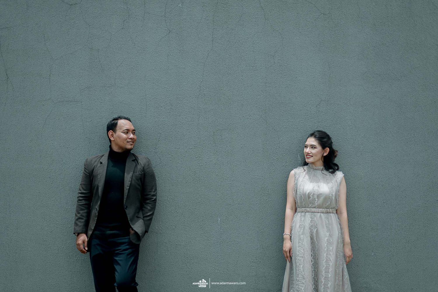 A couple stands against a grey wall in contrasting formal outfits.