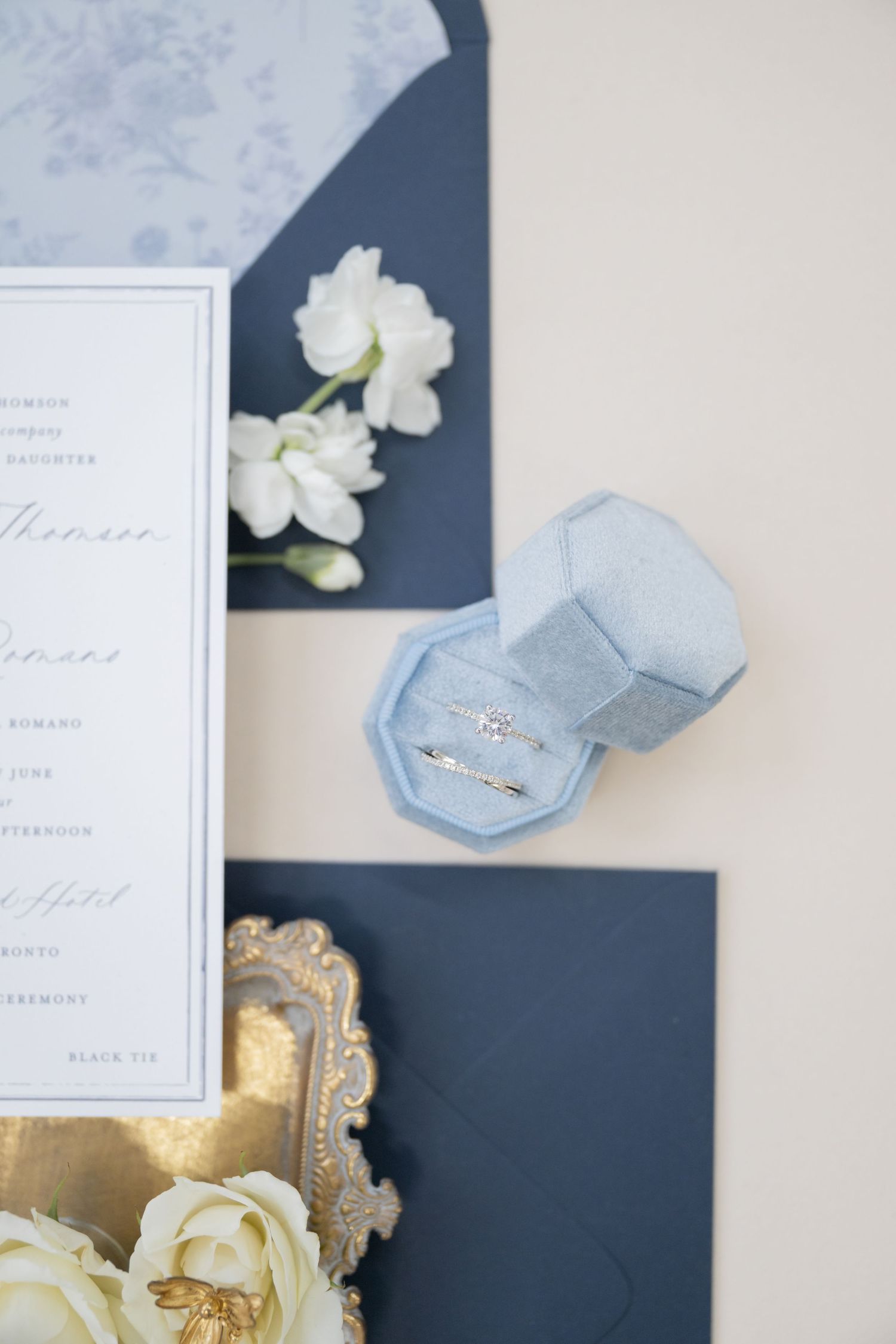 Light blue velvet ring box with wedding stationery and white roses on neutral background.