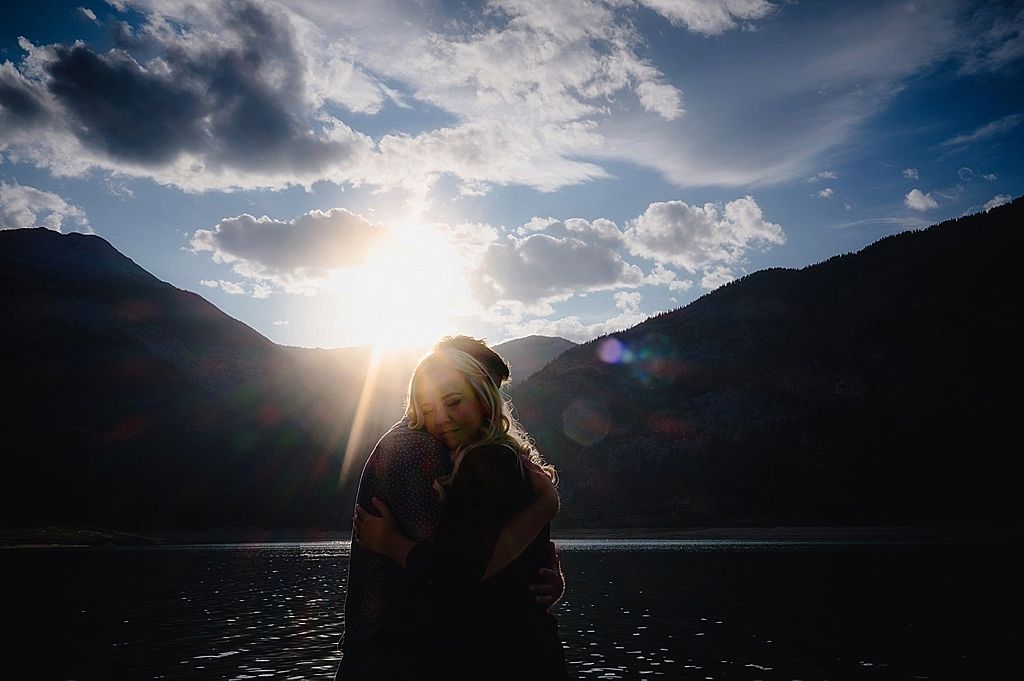 Silhouette of figures by a lake at sunset with dramatic cloud formations and mountain peaks.