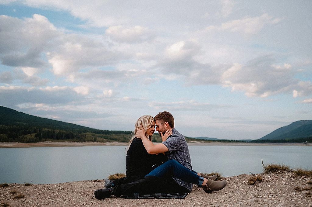 Couple sits close together on rocky shoreline with lake and mountains in background.