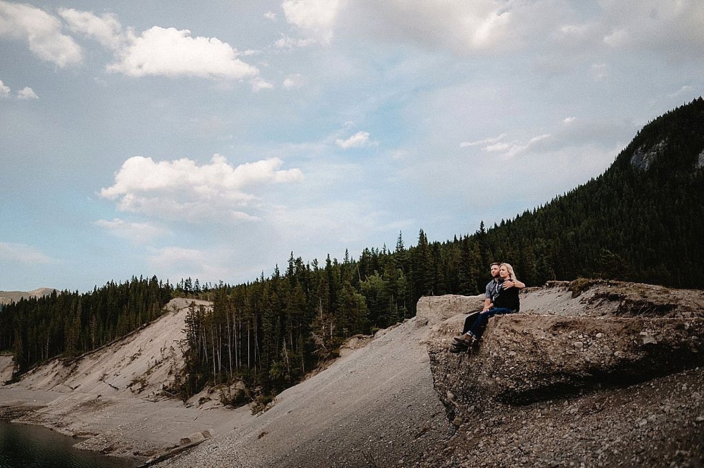 Figure sits on rocky mountain overlook with pine forest and valley views.