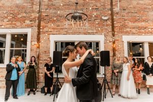 Wedding couple's first dance in front of exposed brick wall with string lights and guests watching.