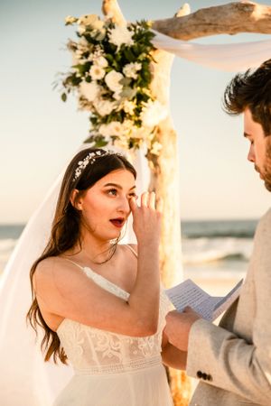 An emotional beach wedding ceremony moment under a floral arch at sunset.