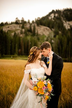 A romantic wedding couple embraces in a mountain meadow with golden grass and evergreen forest backdrop.